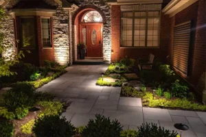 Beautifully lit home entrance with landscape lighting highlighting the pathway, plants, and brick exterior at night.