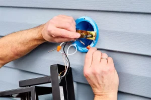 Electrician repairing outdoor landscape lighting wiring on house siding to prevent fixture damage in Raleigh, NC.