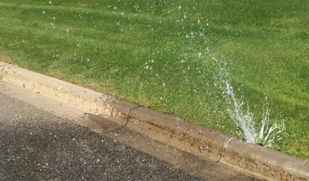Leaking irrigation system with water spraying onto the street, indicating a malfunction near the curb