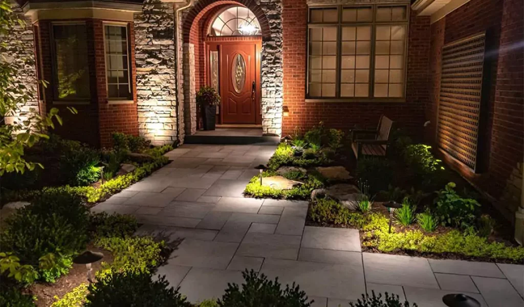 Beautifully lit home entrance with landscape lighting highlighting the pathway, plants, and brick exterior at night.